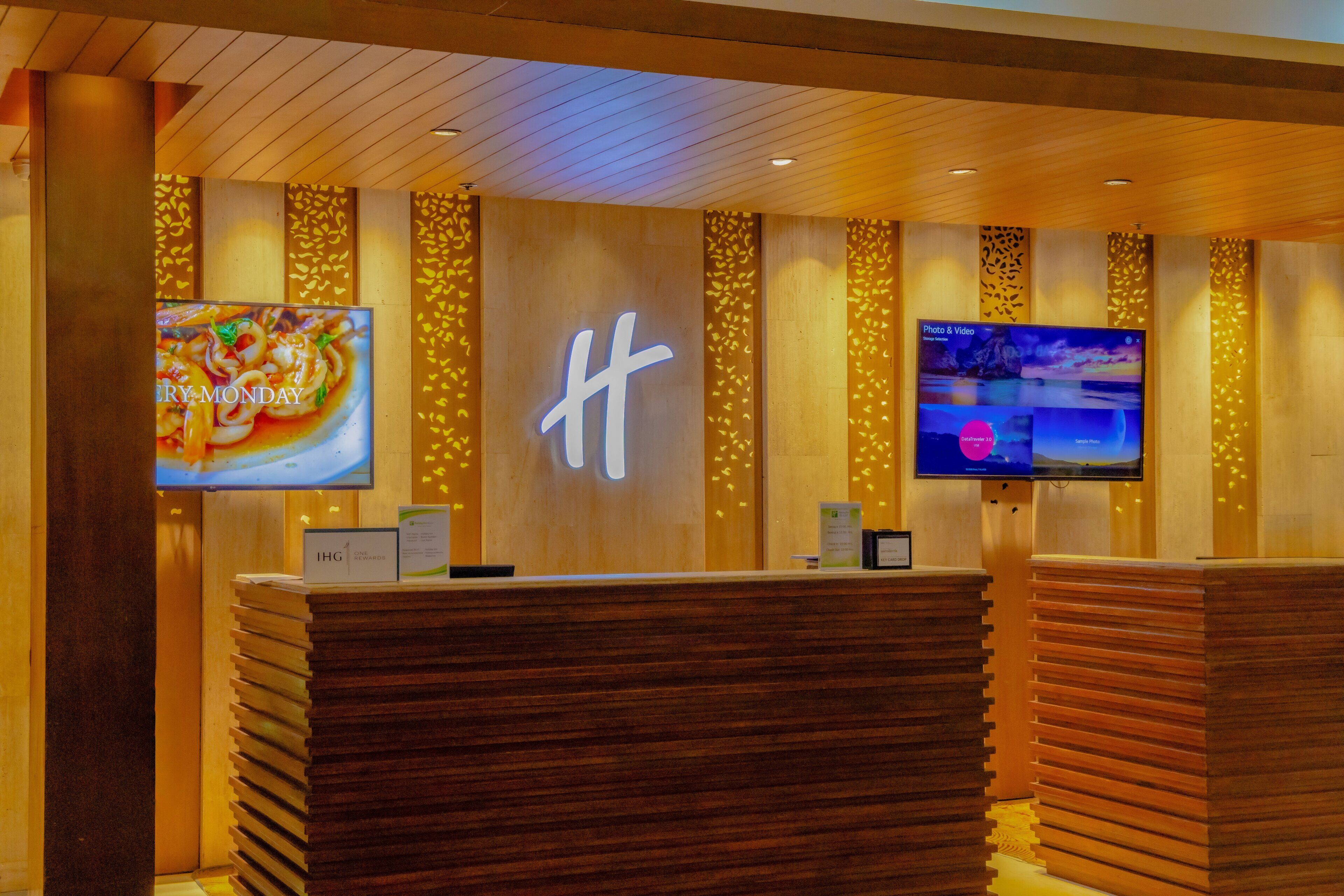 Zona de recepción de hotel con paneles de madera, dos televisiones de pantalla plana grandes montadas en la pared y un mostrador de recepción con una pila de folletos.