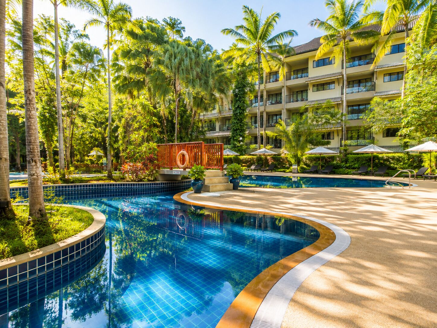 A resort pool area with a curved pool, palm trees, and a multi-story building with balconies.