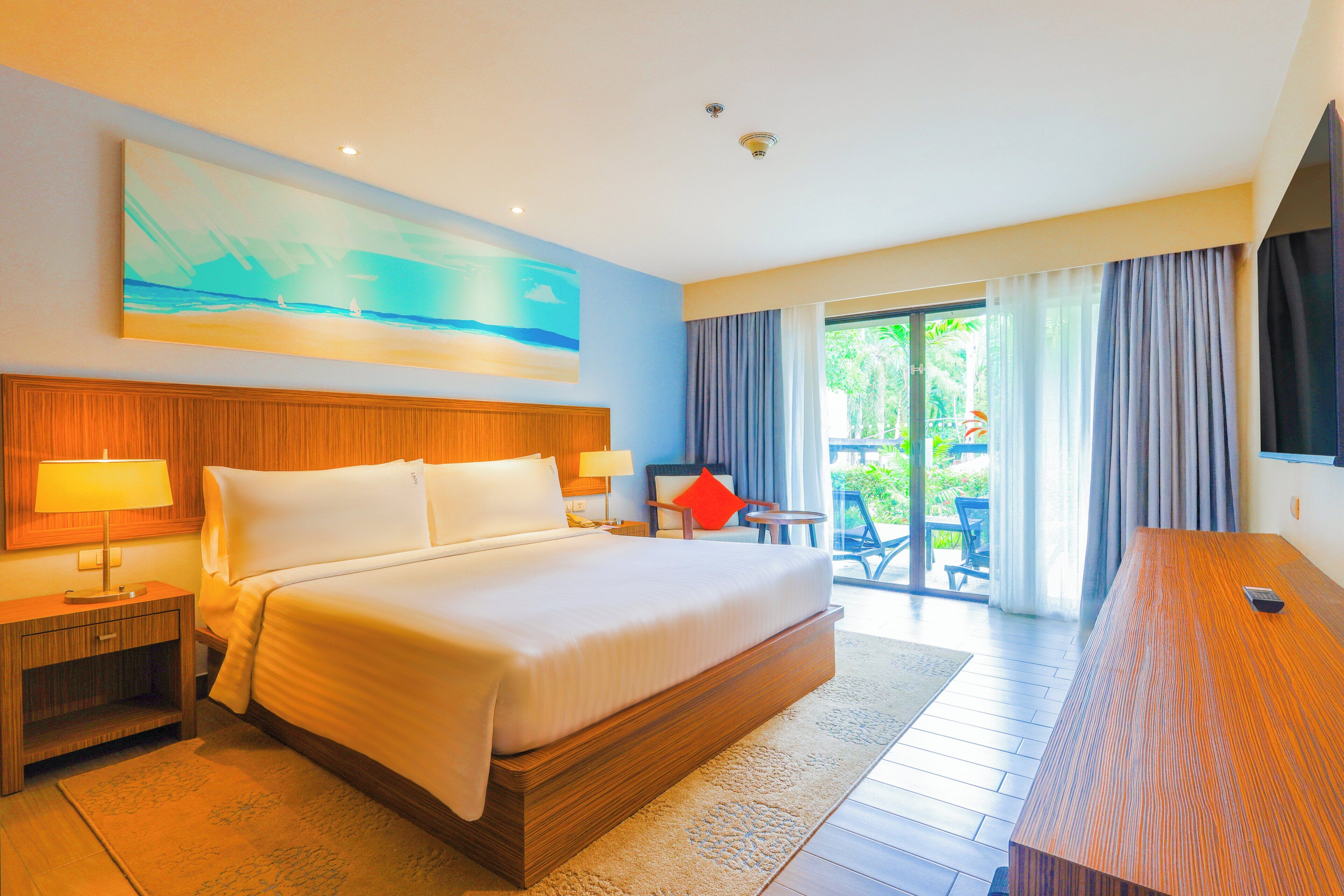 A hotel room with a large bed, a wooden headboard, a nightstand, a desk, and a view of the outdoors through a glass door.