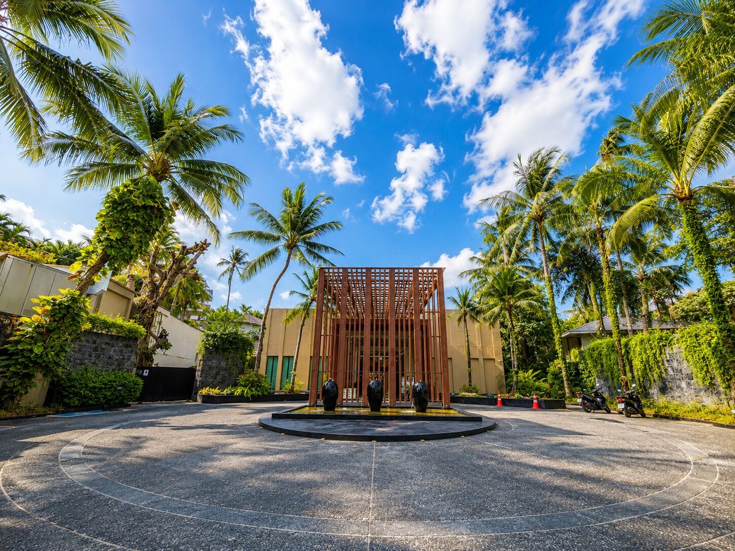 A circular driveway leading to a building with a wooden fence structure, surrounded by palm trees and a clear sky.