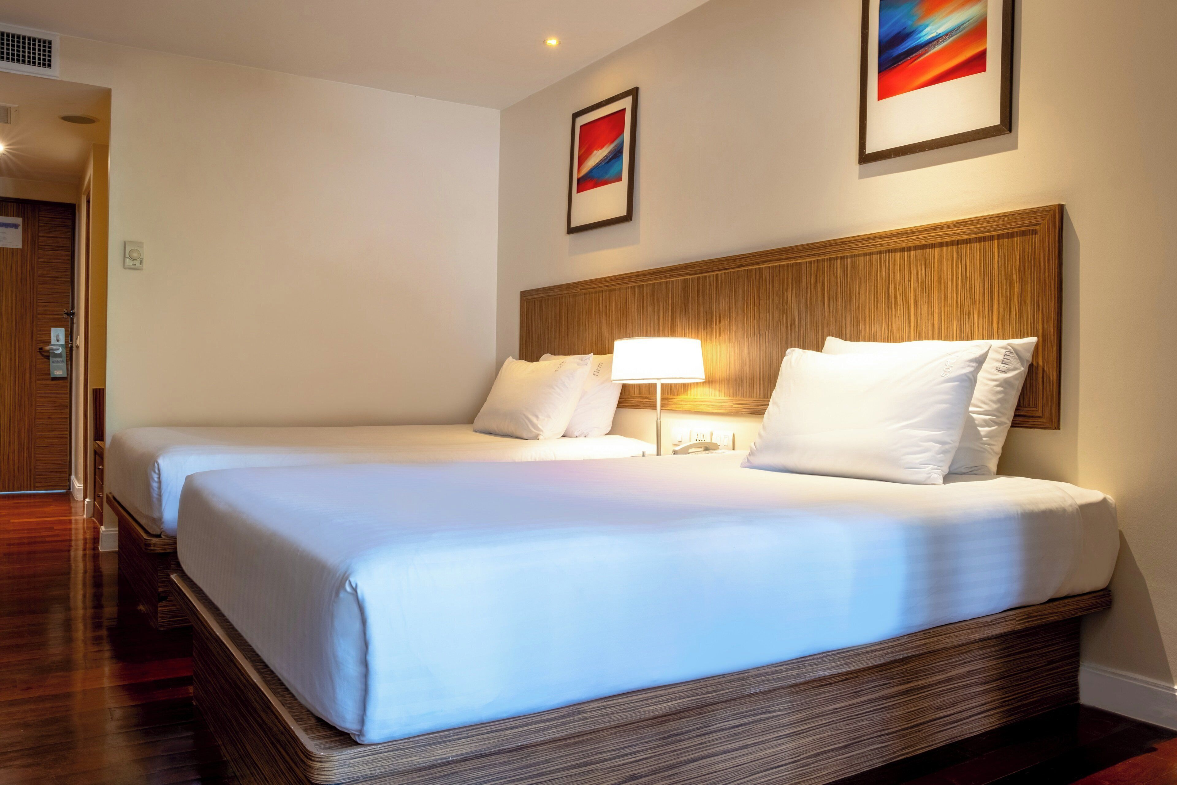 A hotel room with two beds, a wooden headboard, a bedside lamp, and two framed pictures on the wall.