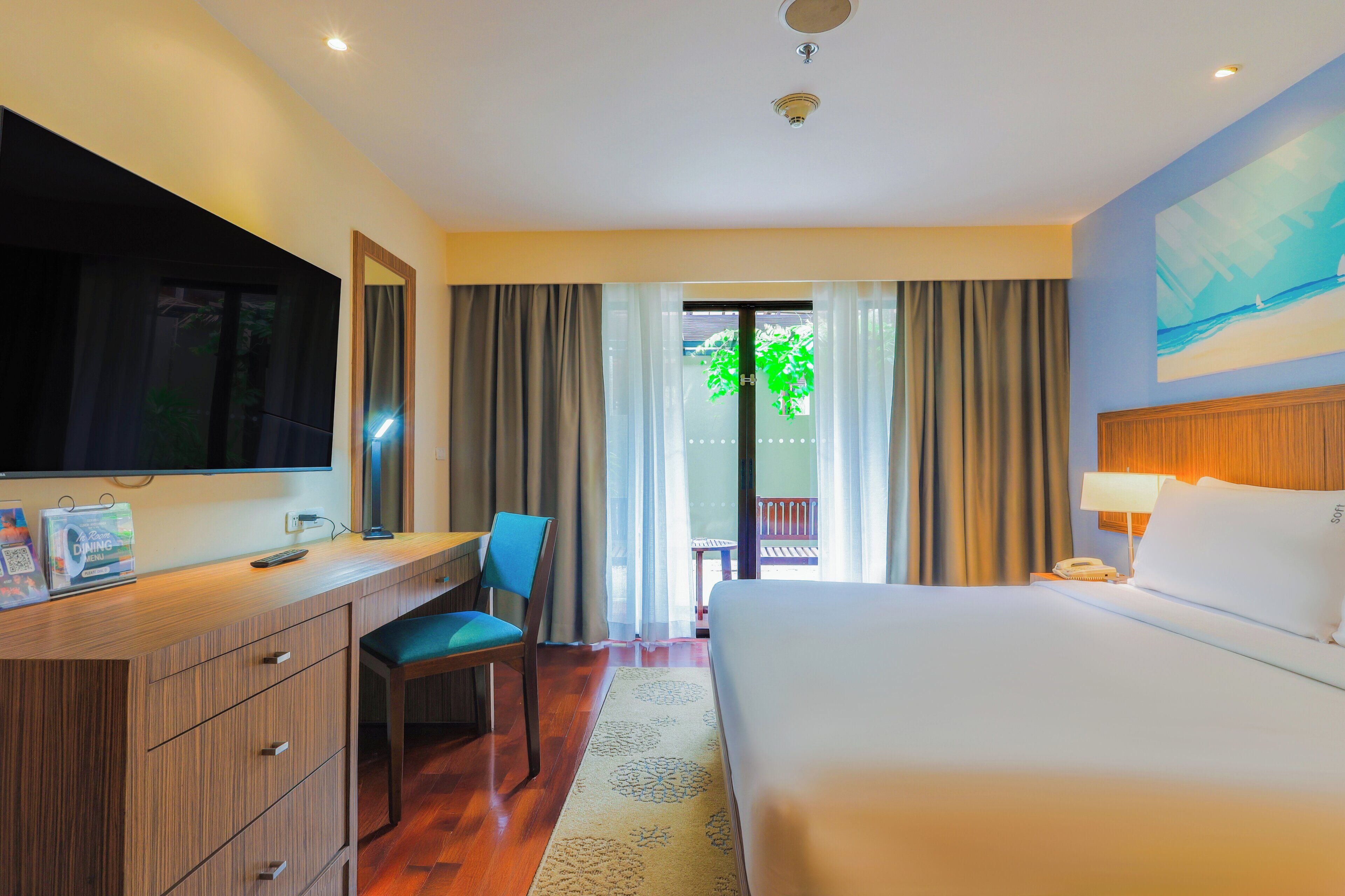 A hotel room with a bed, desk, chair, TV, and a view of greenery outside.