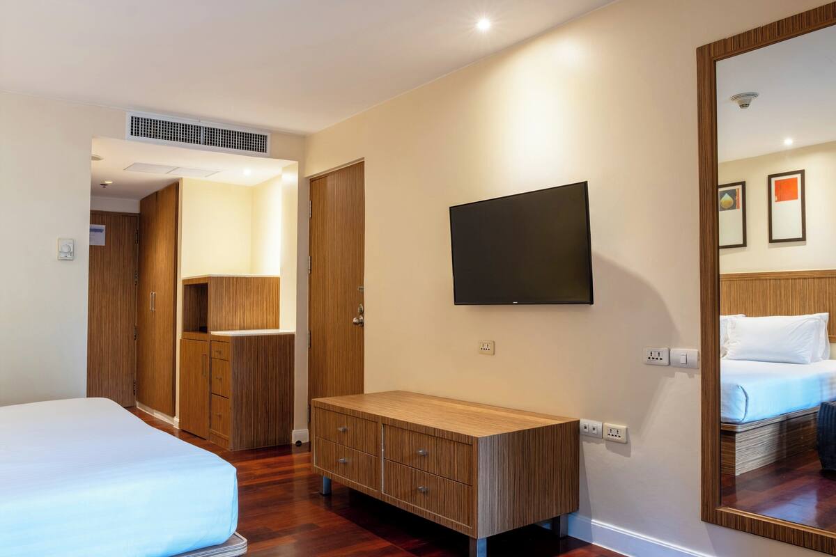 A hotel room with a bed, a wooden dresser, a television mounted on the wall, and a mirror reflecting the bed.