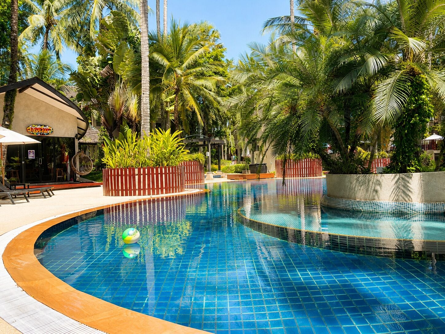 A swimming pool with a tiled edge, surrounded by palm trees and a building with a sign.