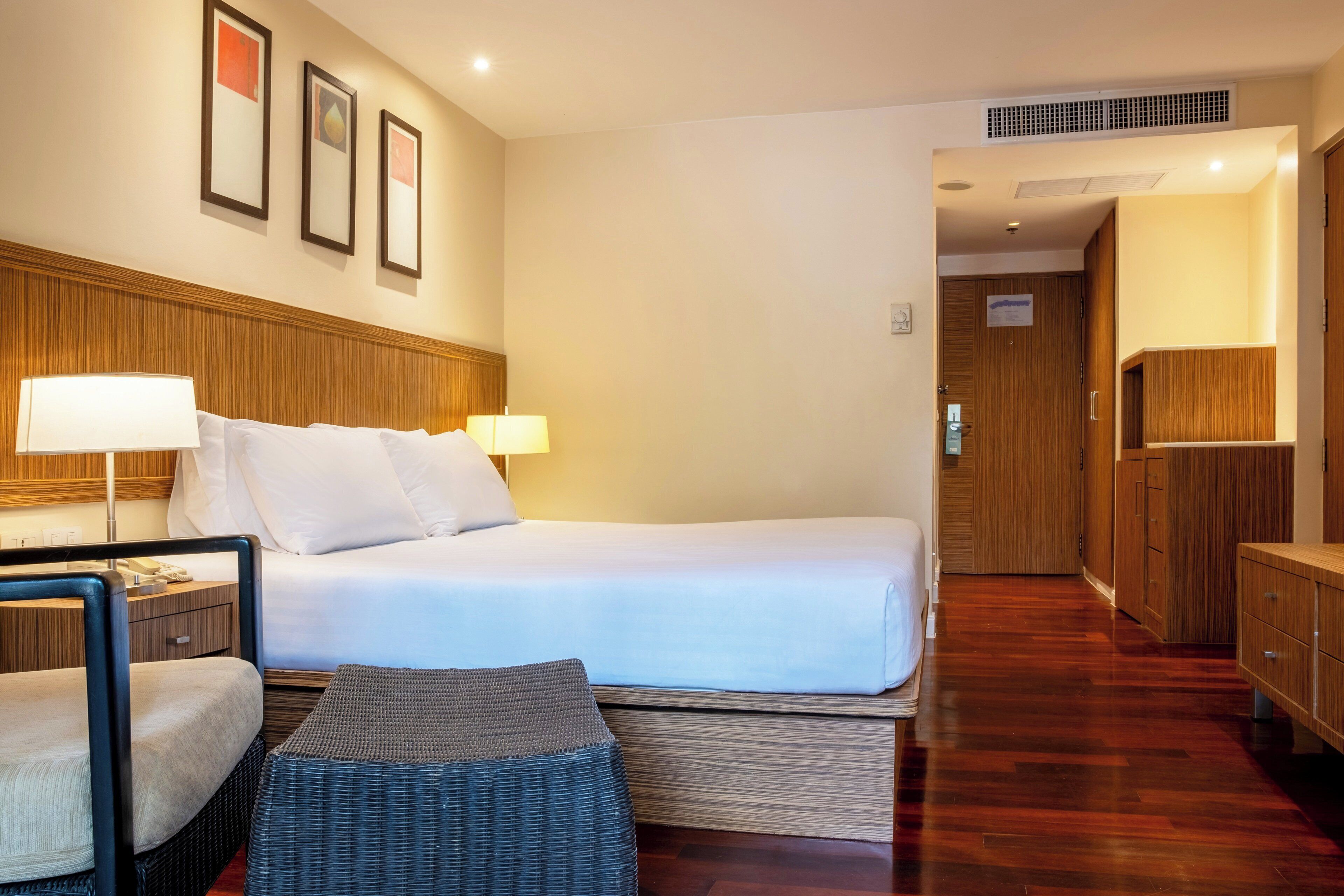 A hotel room with a large bed, two bedside lamps, a wooden headboard, a wicker chair, and a wooden wardrobe.