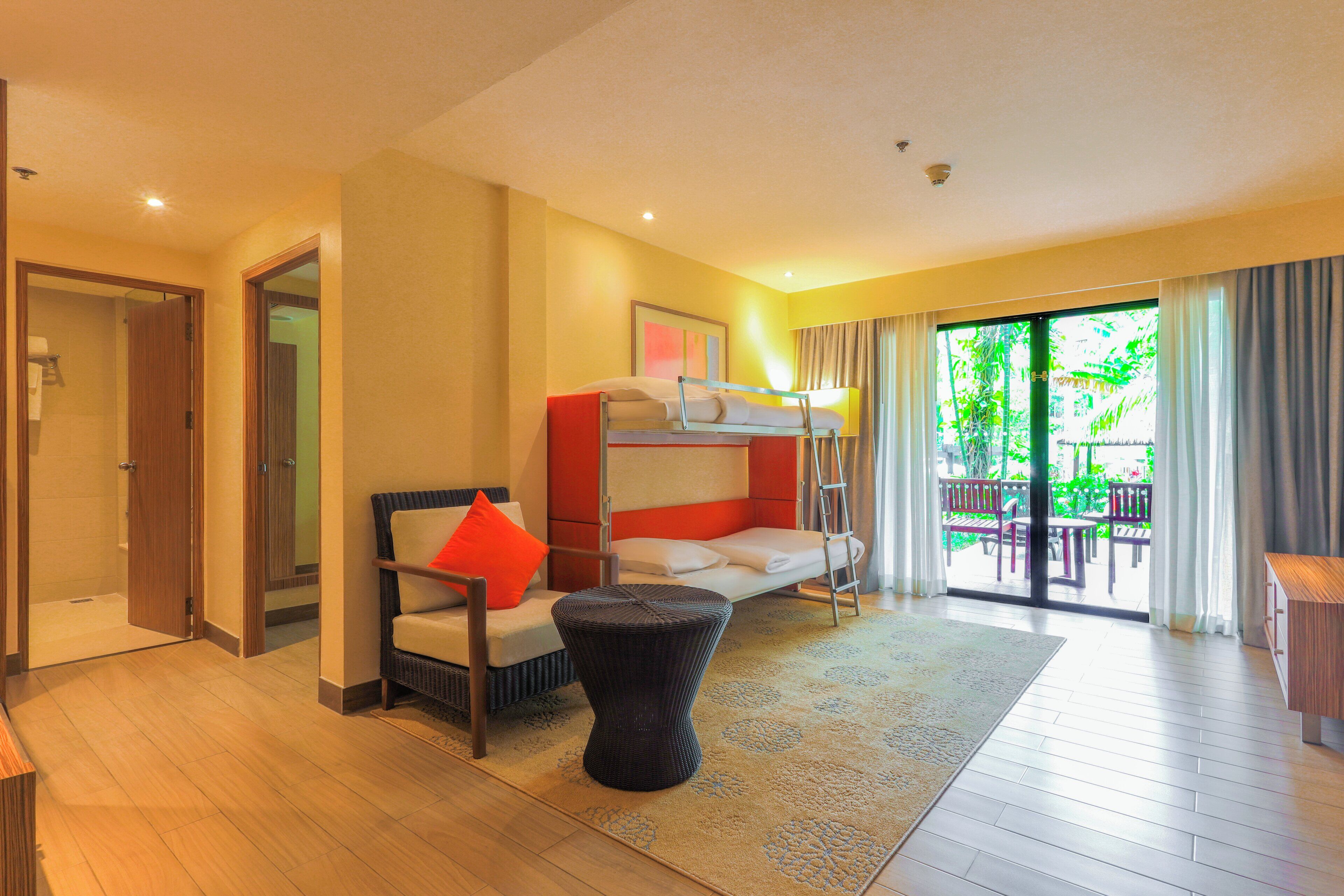 Habitación de hotel con literas, una silla con almohada roja, una mesita redonda y vistas al exterior a través de una puerta corrediza de vidrio.
