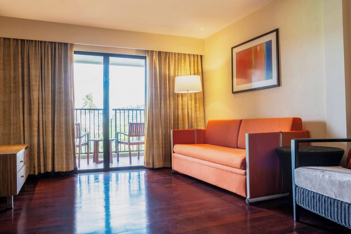 A hotel room with a wooden floor, a sofa, a chair, a table, a lamp, and a balcony with a view.