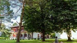 A building with a two-story balcony, surrounded by a well-maintained lawn and trees. - Regina Hotel (Druskininkai)