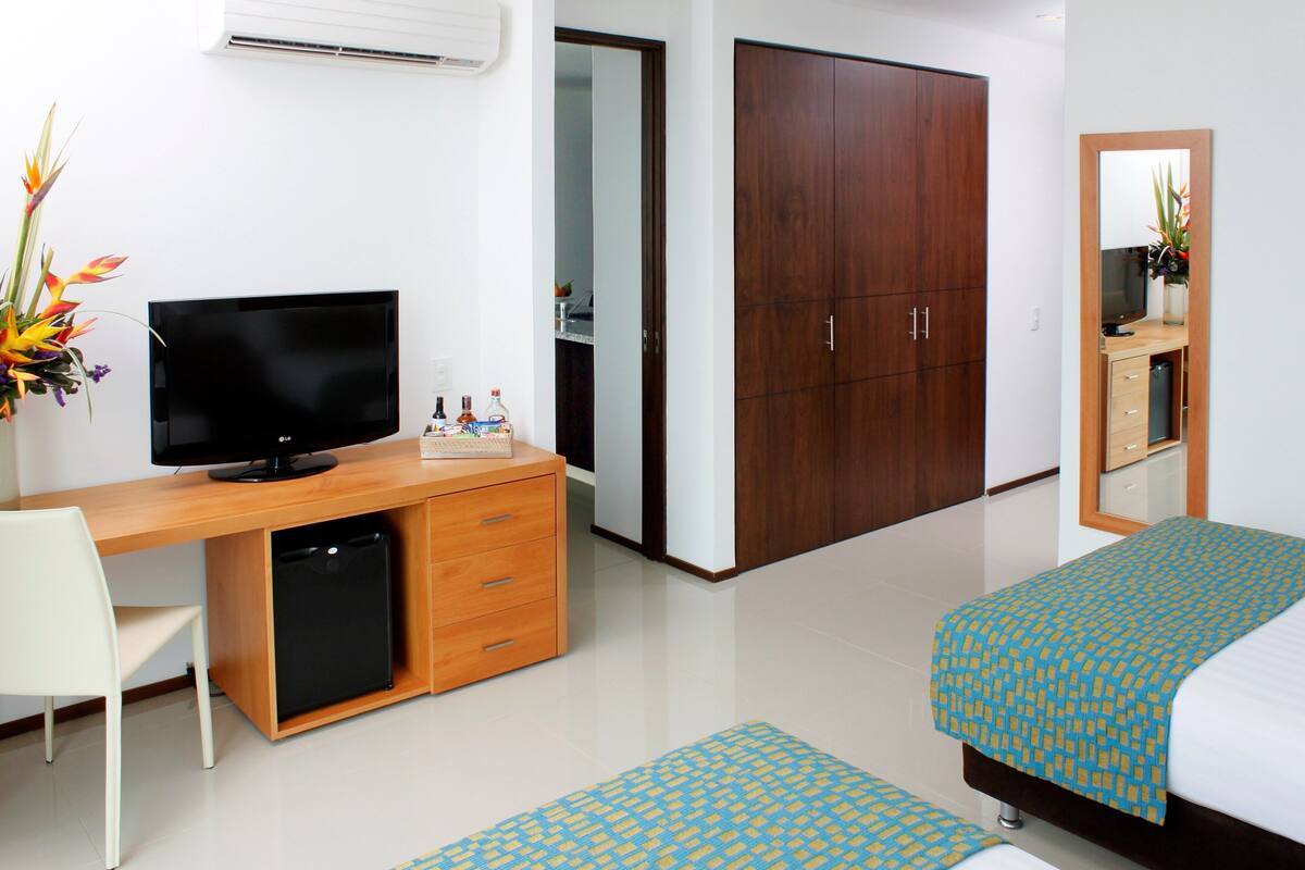 Habitación de hotel con un escritorio de madera, televisión, armario, cama con colcha estampada y un refrigerador pequeño.
