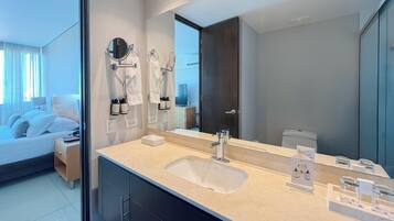 A modern hotel bathroom with a large mirror, a sink, and a toilet. There is a bed with a nightstand and a lamp in the adjacent room.