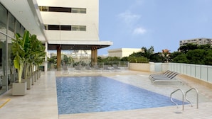 Una piscina al aire libre con sillones de descanso y plantas en macetas, rodeada por un edificio moderno con amplias ventanas.