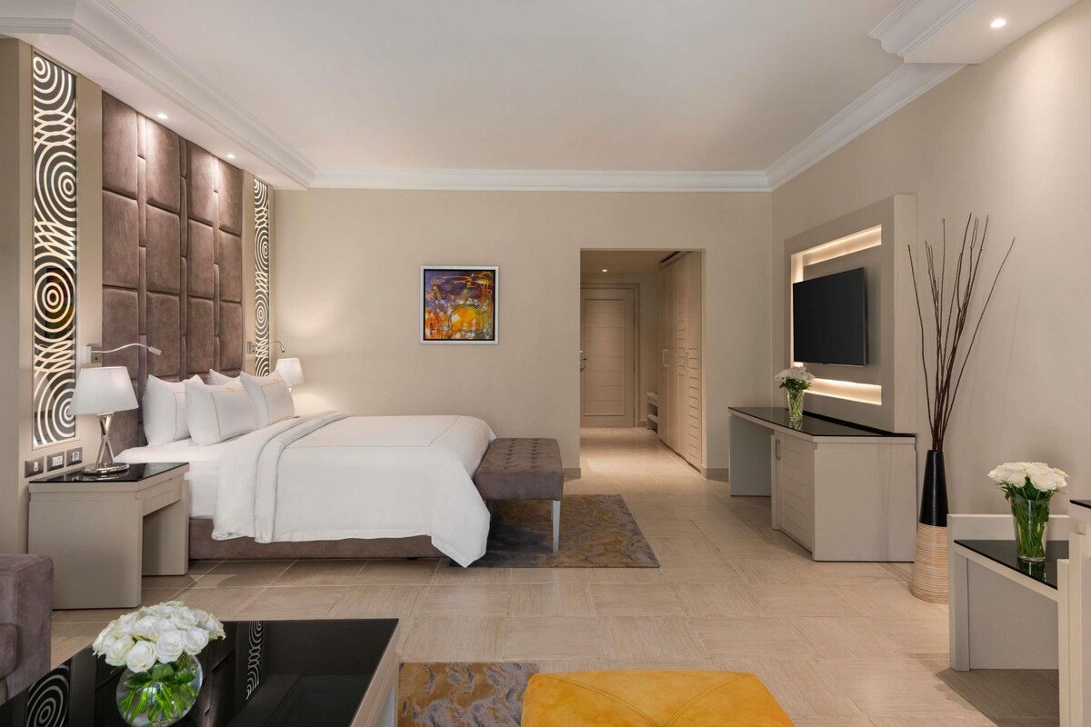 A modern hotel room with a large bed, a desk, a TV, and a decorative wall panel.