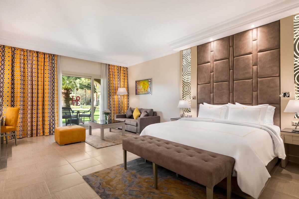 A hotel room with a large bed, a sofa, a bench, and a dining area with a table and chairs.