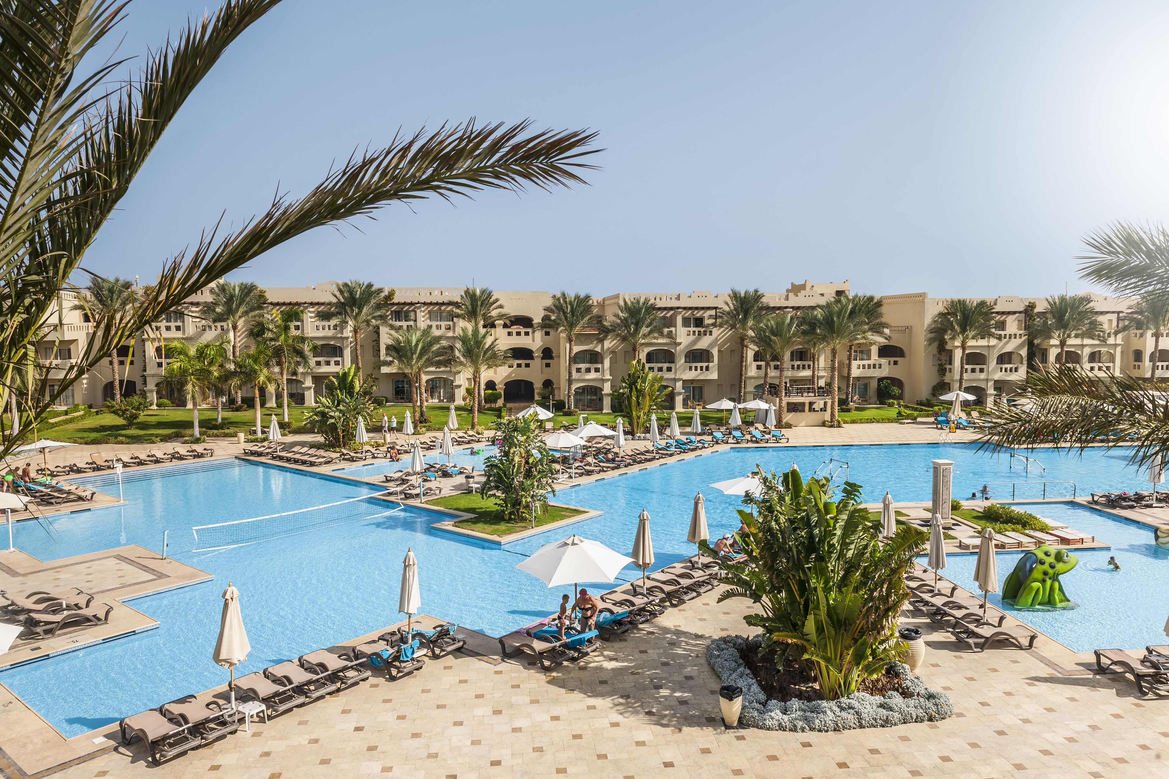 A large pool area with multiple swimming pools, sun loungers, umbrellas, and palm trees.