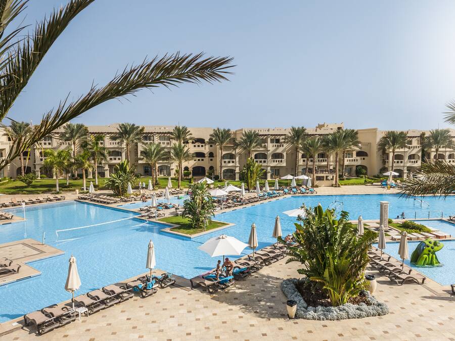 A large pool area with multiple swimming pools, sun loungers, umbrellas, and palm trees.