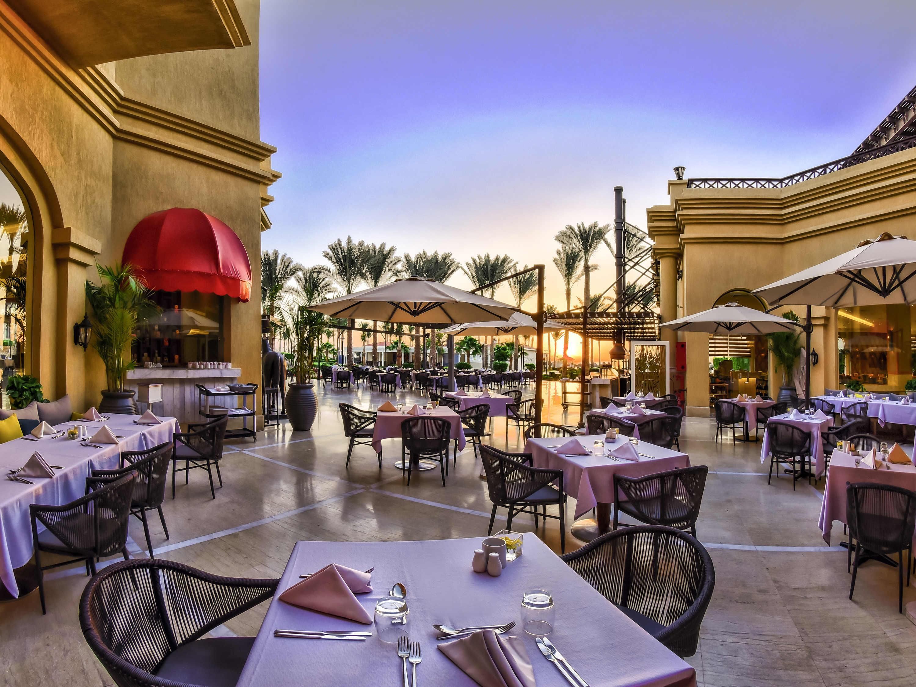 An outdoor dining area with tables and chairs, umbrellas, and palm trees.