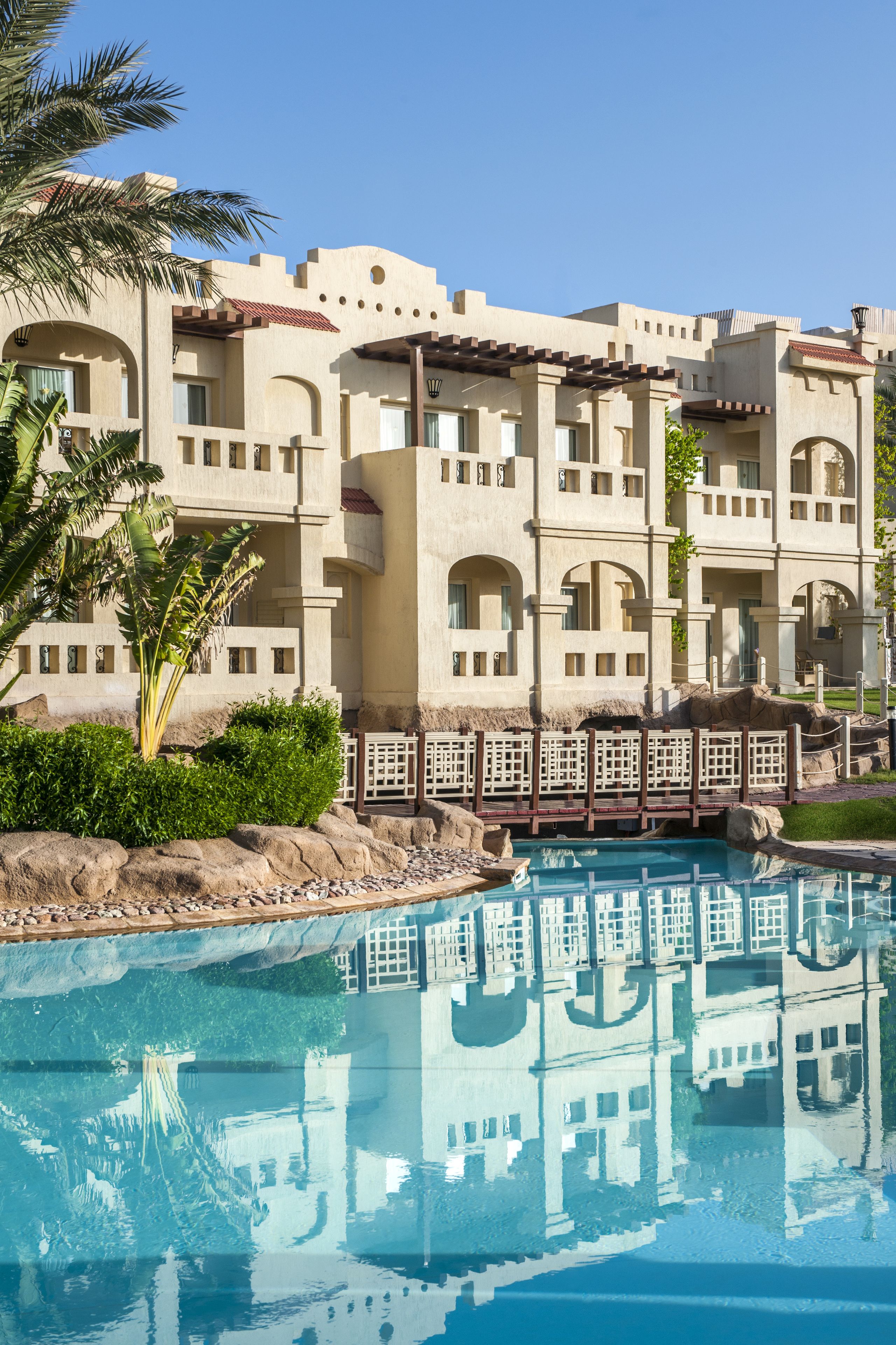 A hotel with a pool, palm trees, and a reflection of the building in the water.