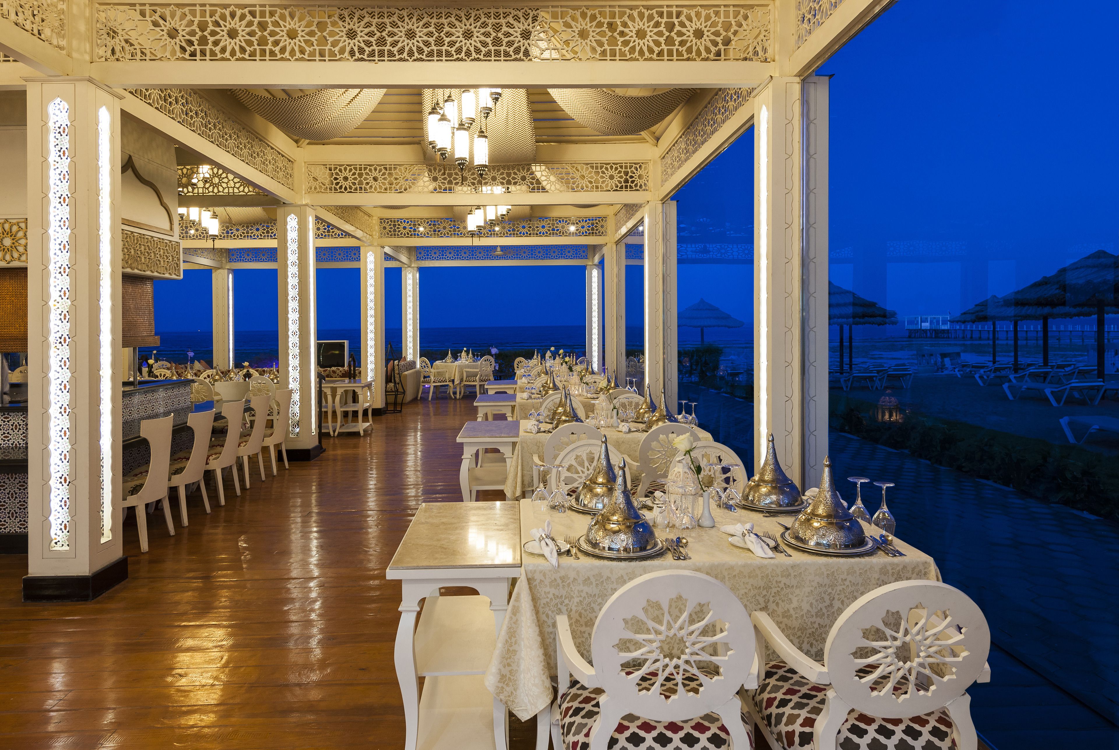 Een restaurant met witte meubels en gedetailleerde plafonddecoraties, met uitzicht op een waterpartij.