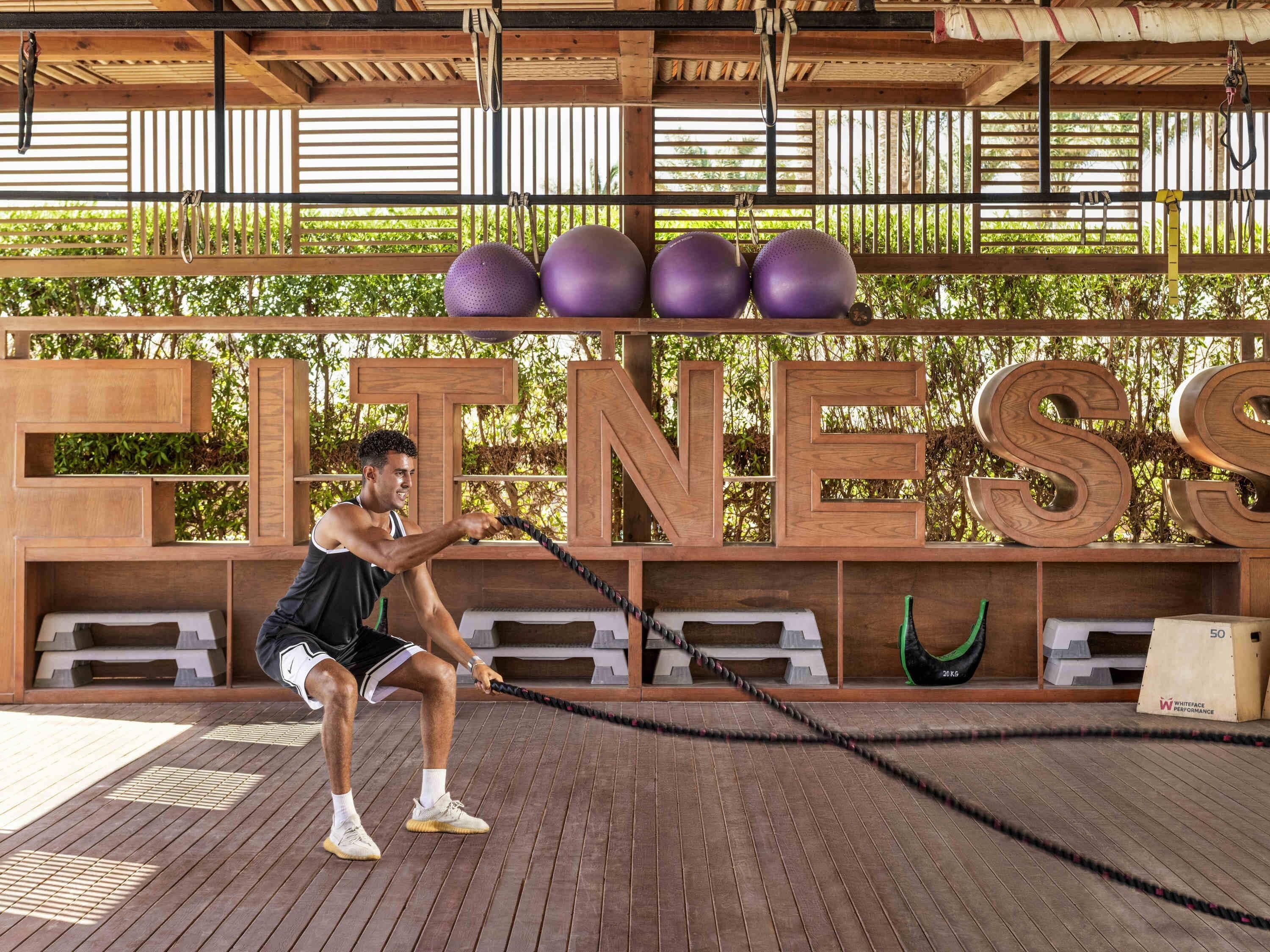 A man performing a workout with a battle rope in a wooden-floored area with a 'FITNESS' sign in the background.