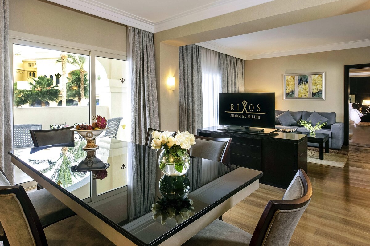 A modern living room with a dining area, a flat-screen TV, and a view of palm trees.