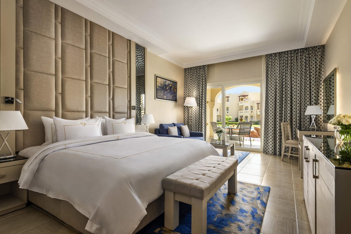 A hotel room with a large bed, a desk, a chair, a bench, and a view of the outside.