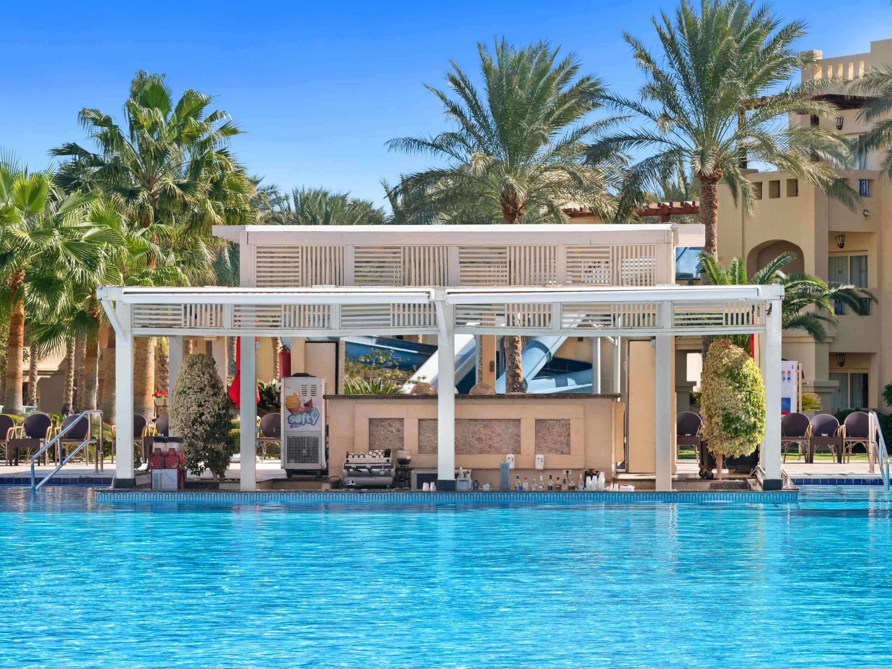 A pool area with a covered seating area, palm trees, and a building in the background.