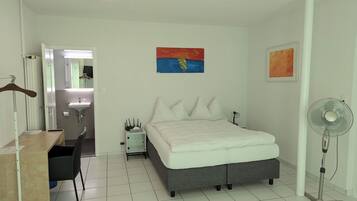 Une chambre à coucher comprenant un lit, un bureau, une chaise, une table de chevet, un miroir, un ventilateur fixé au mur et un tableau.