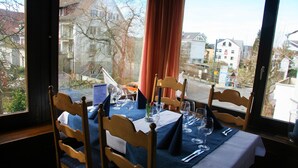 A dining table set for four with wooden chairs, a window view of a residential area, and a red curtain partially drawn. - Hotel Eremitage (Arlesheim)