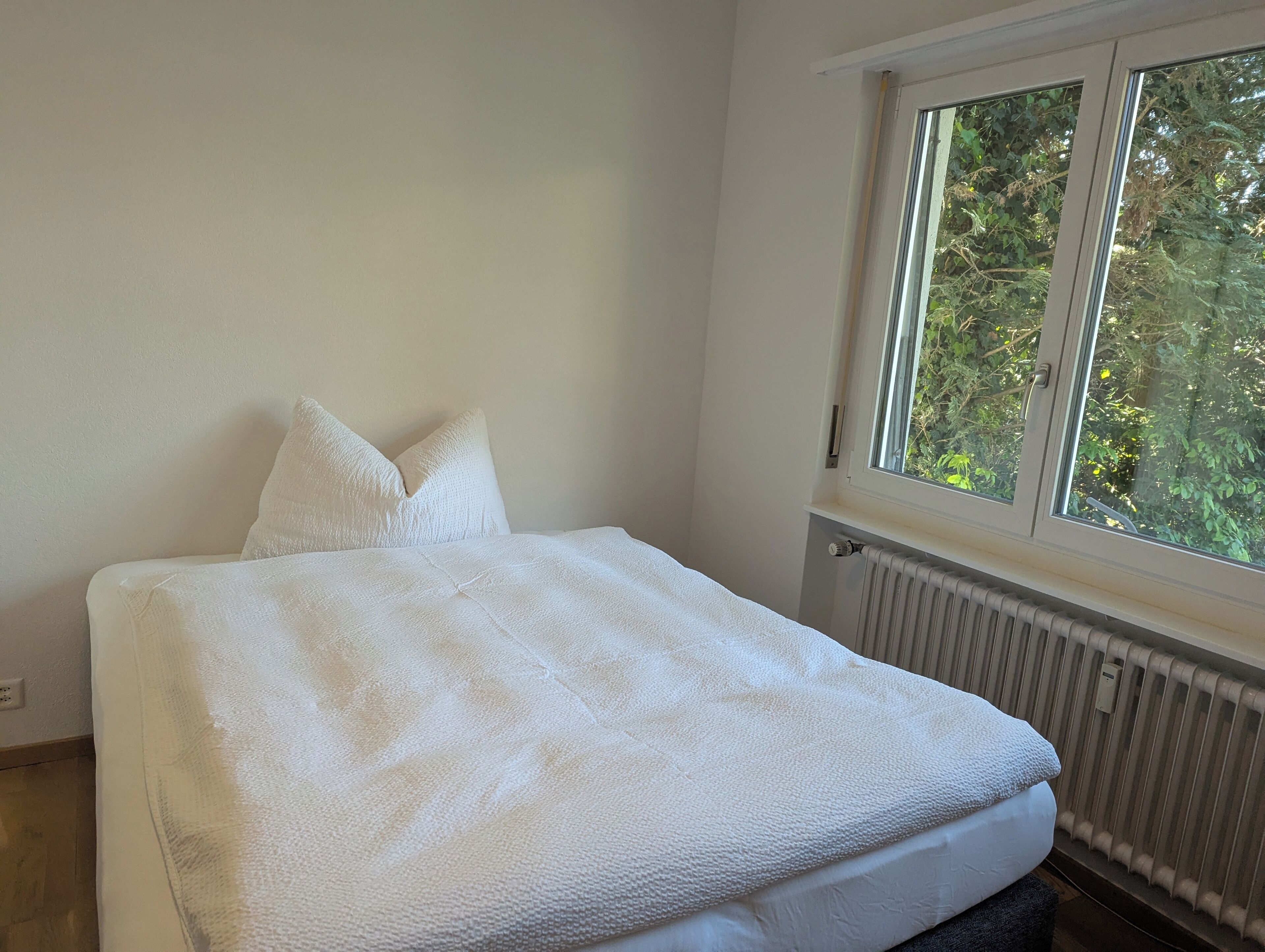 Ein Schlafzimmer mit einem Bett, weißen Kissen, einem Fenster und einer Heizung.