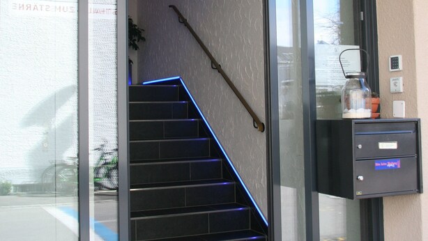 A hotel entrance with a glass door, a staircase with blue lighting, and a small desk with a coffee machine. - Hotel Eremitage (Arlesheim)