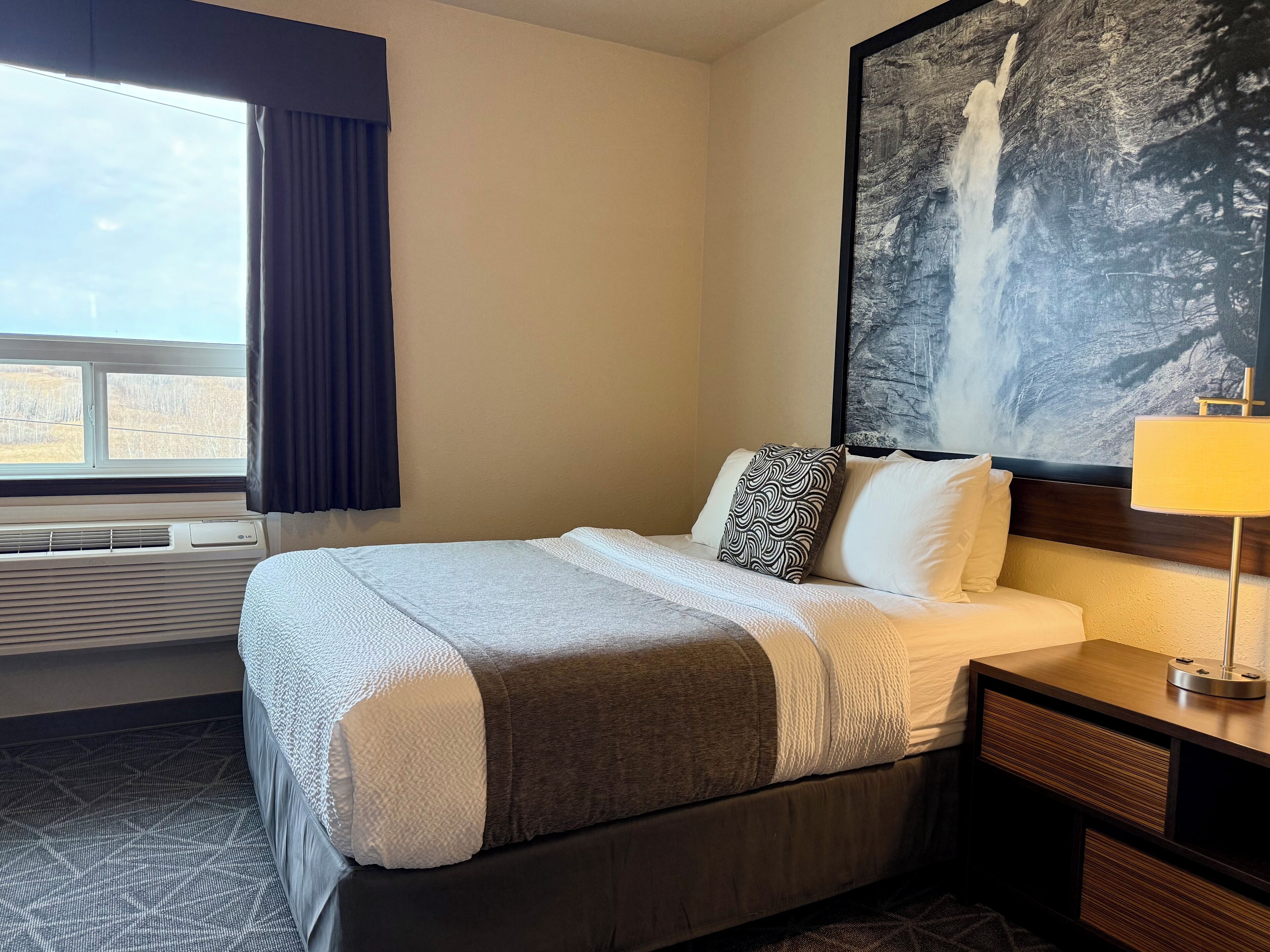 Hotelkamer met een bed, nachtkastje, lamp, groot schilderij van een waterval, raam met gordijnen en een airconditioning.