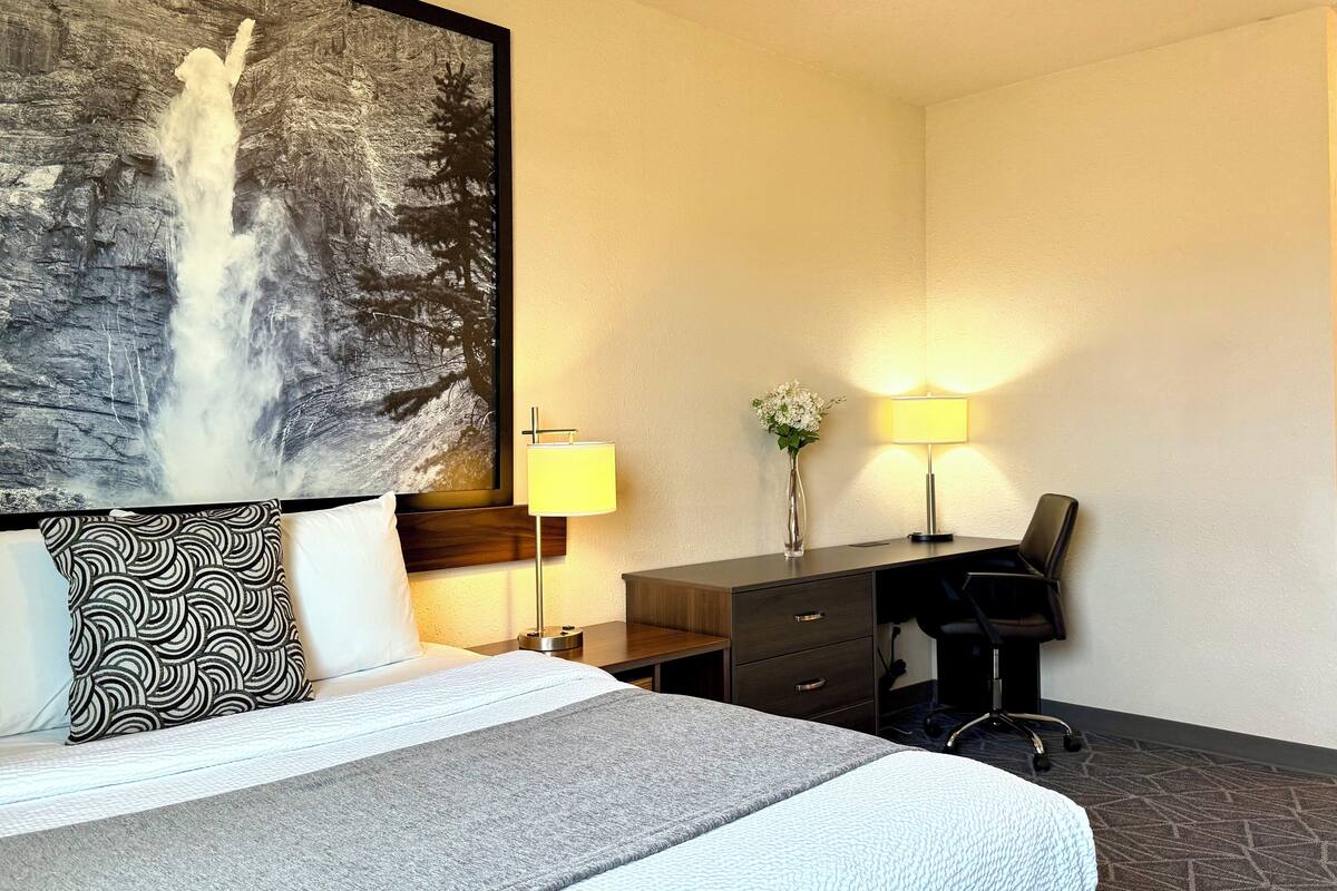 A hotel room with a bed, a desk, a chair, a lamp, a vase with flowers, and a large framed picture of a waterfall.