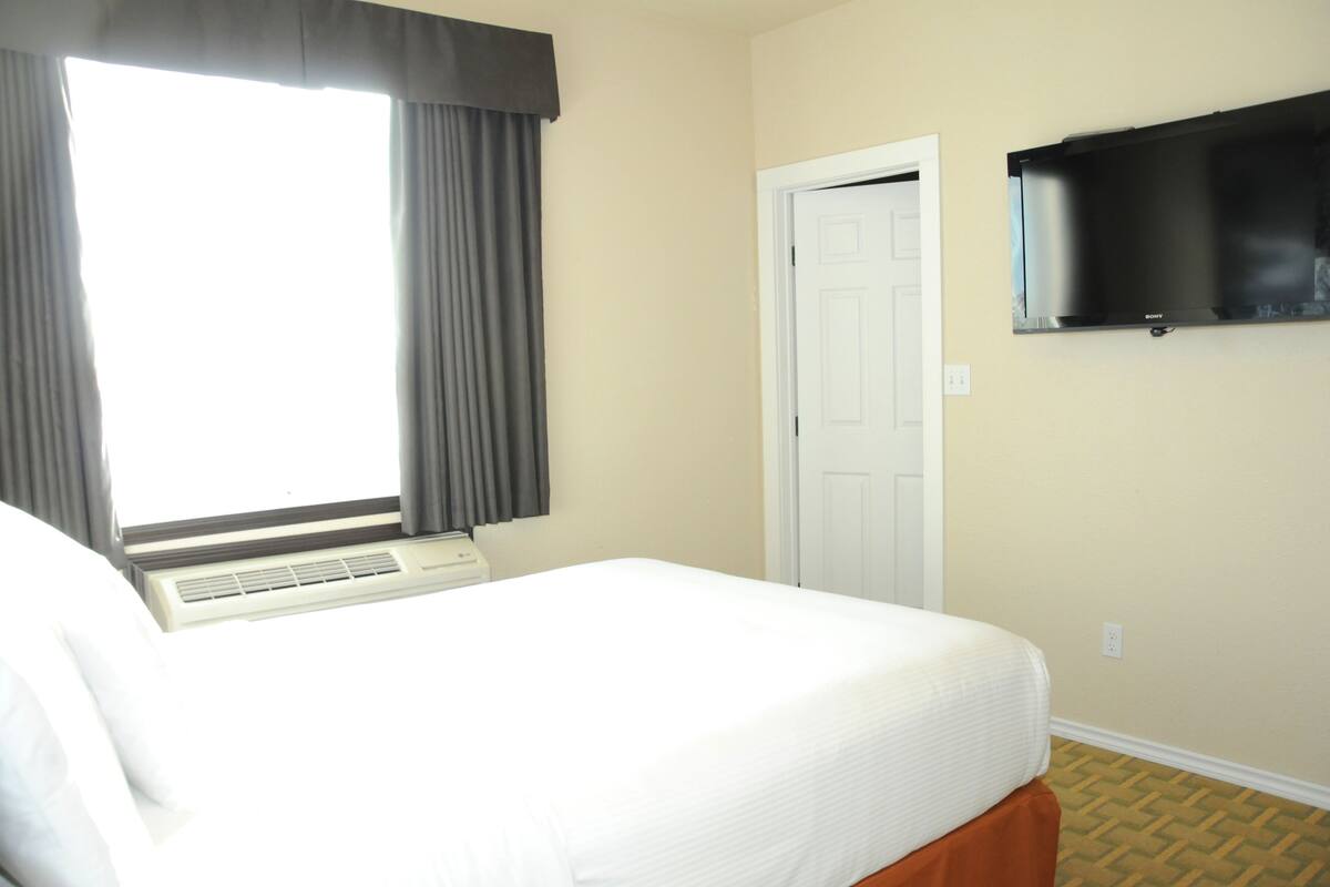 A hotel room with a bed, a window with curtains, a door, and a mounted television.