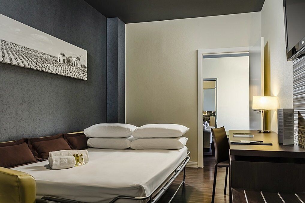 Une chambre d’hôtel avec un lit, un bureau et une chaise. Il y a un tableau au mur et une lampe sur le bureau.