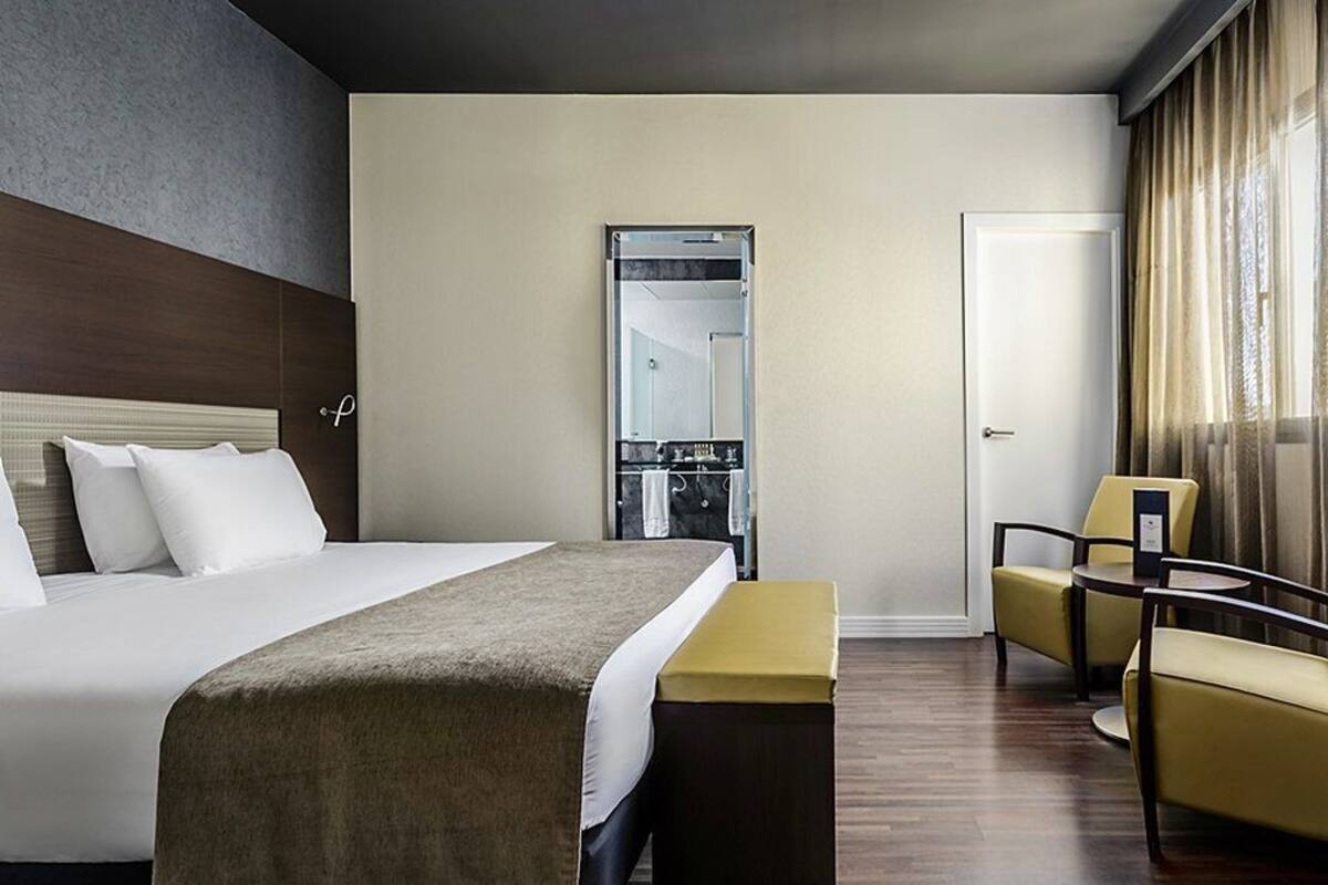 Ein Hotelzimmer mit einem großen Bett, einer Bank, zwei Stühlen, einem Spiegel und einer Tür.