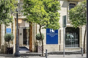 Welcome to Eurostars Asta Regia, where the hotel's entrance features a glass door and a beautiful urban setting with trees and a street lamp.