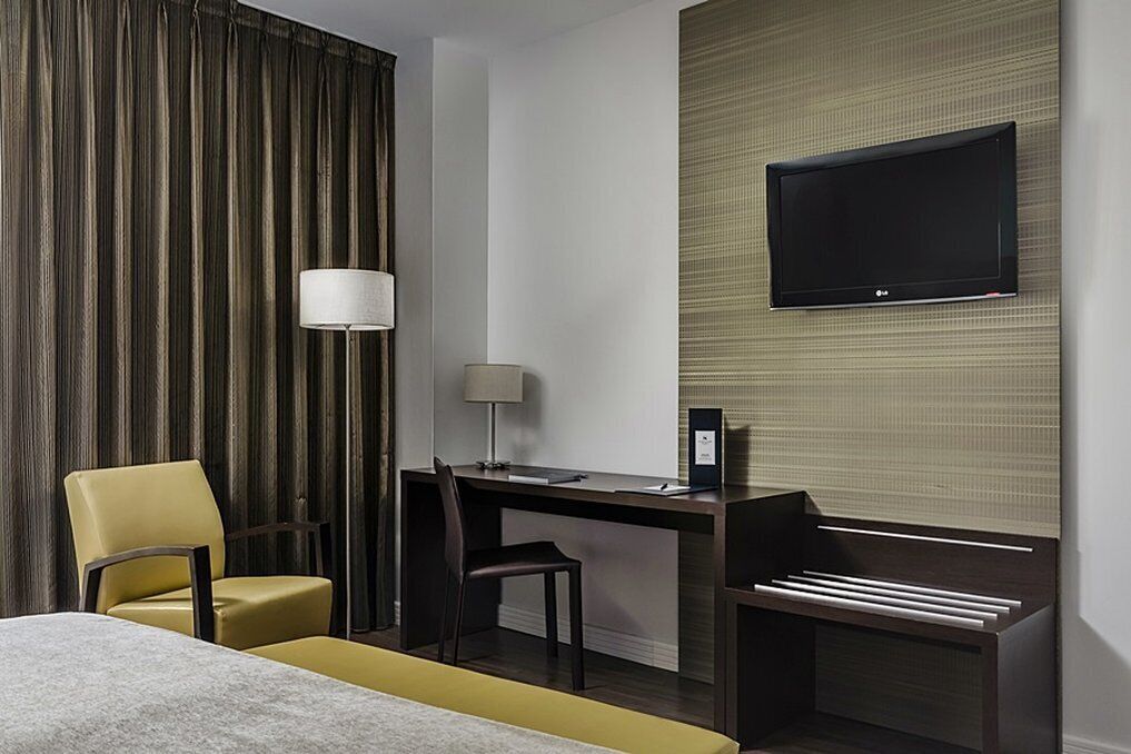 Ein Hotelzimmer mit einem Bett, einem Schreibtisch mit Stuhl, einem an der Wand befestigten Fernseher und einer Lampe.