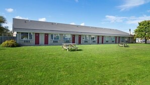 A motel with a row of rooms, each with a window and a door. There are picnic tables in the front lawn.