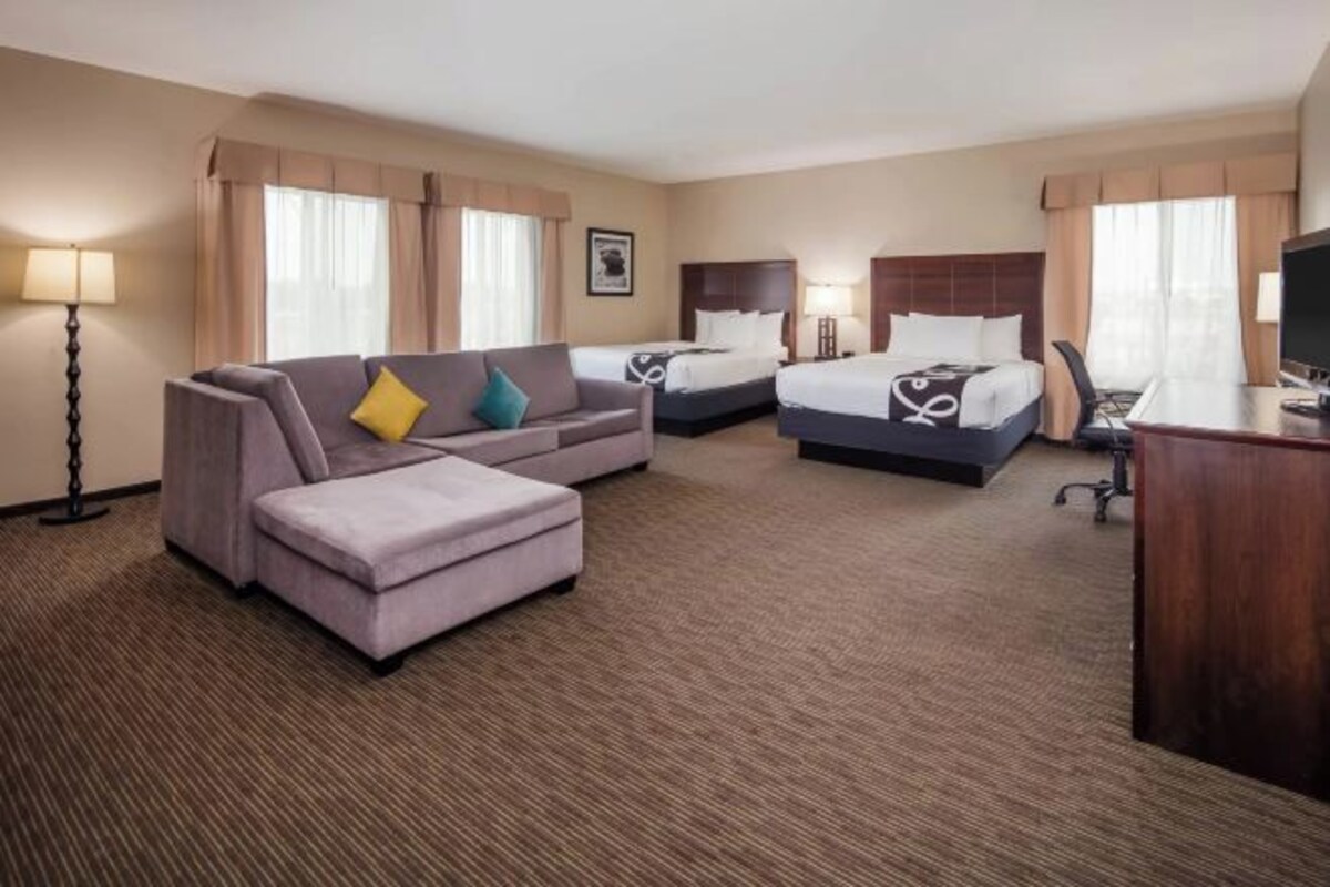 A hotel room with a sofa, two beds, a desk, and a lamp.
