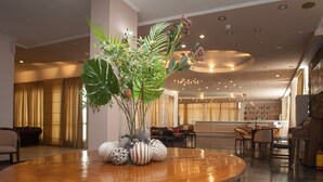 A spacious lobby with a wooden reception desk, a large potted plant, and a seating area with chairs and tables. - Alexandros Hotel -  All Inclusive (Corfu)