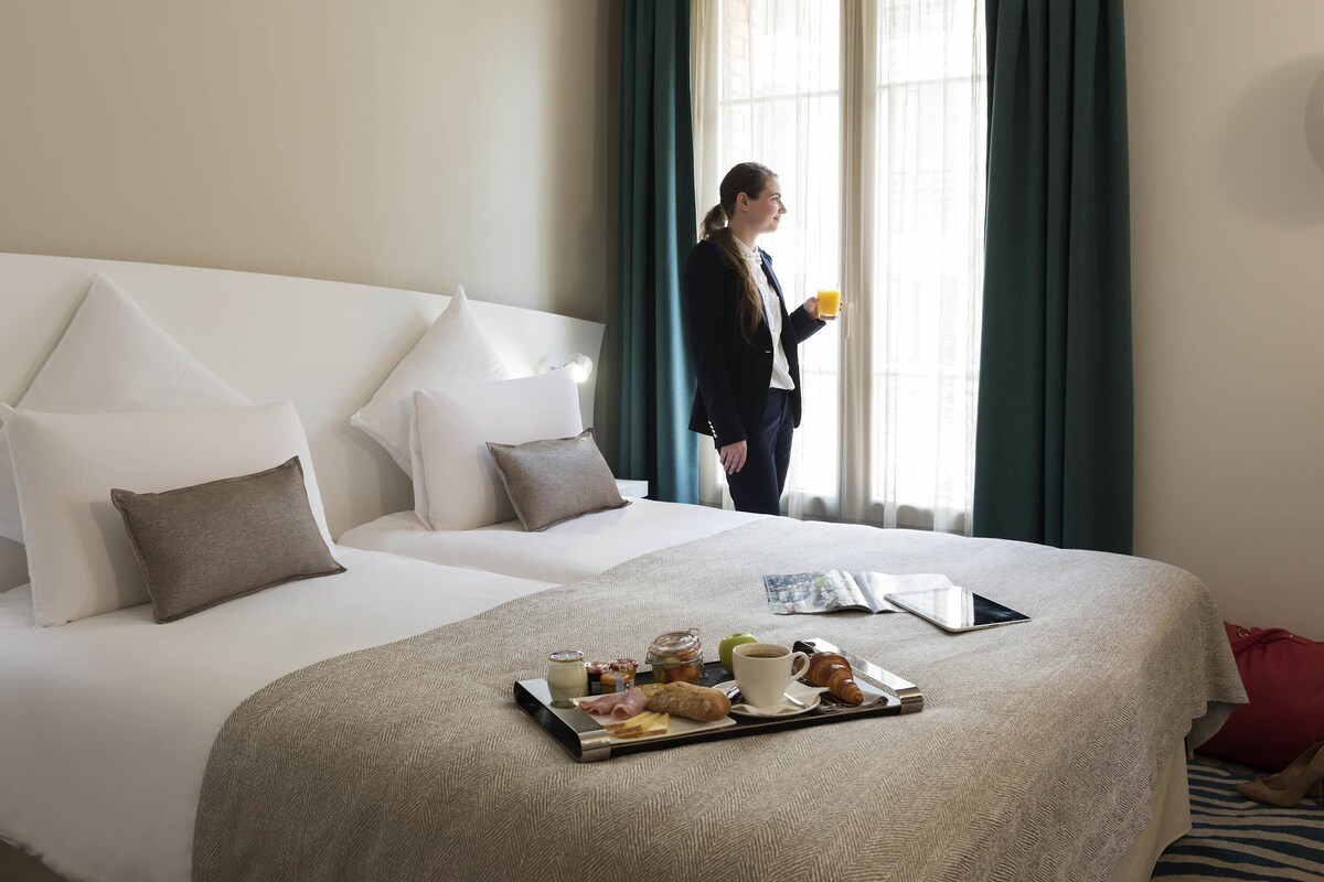 A hotel room with a bed, a tray with breakfast items, and a person standing by the window.