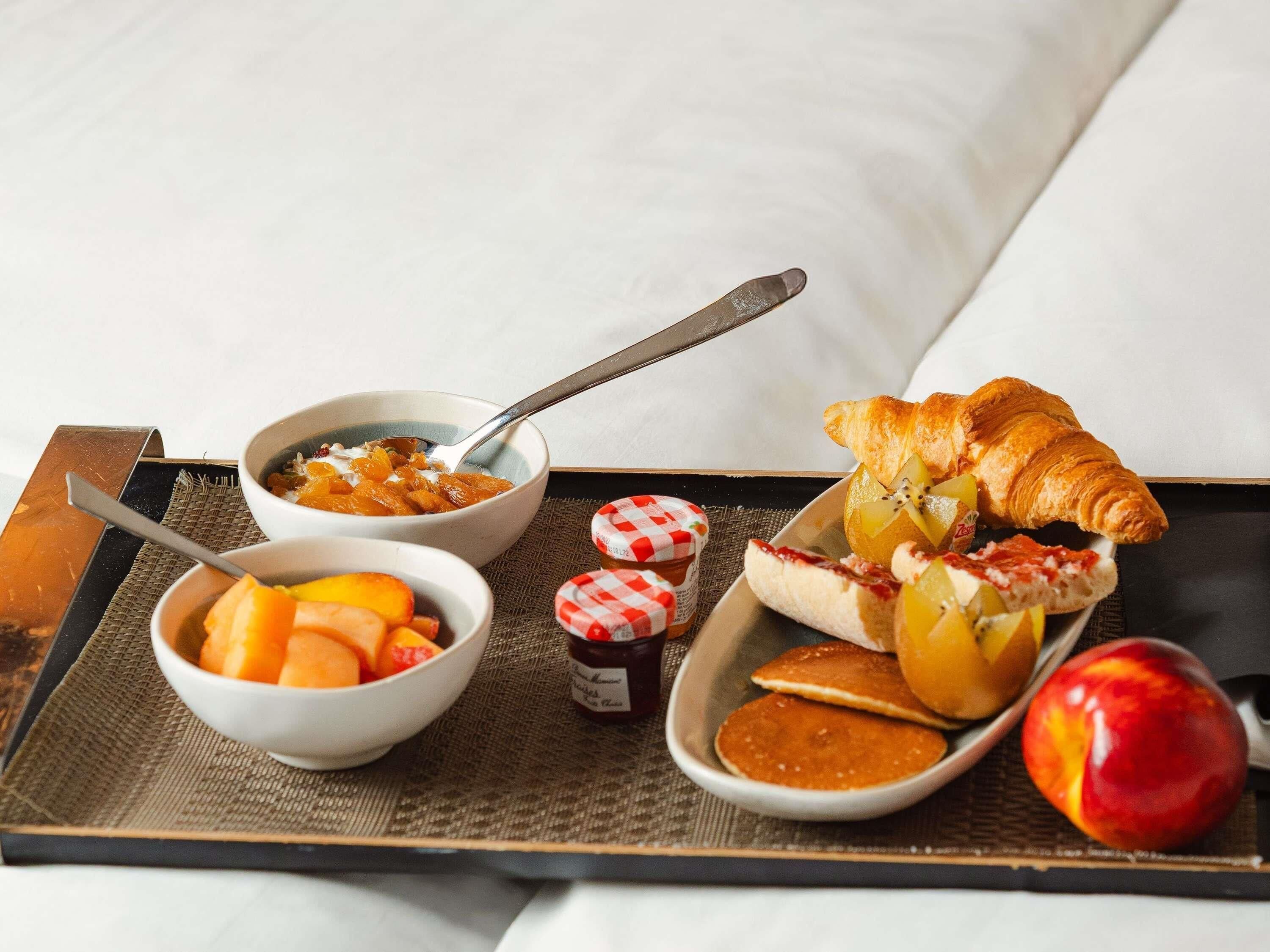 Bandeja de desayuno con un tazón de fruta, un croissant y un pastel con fruta sobre una cama.