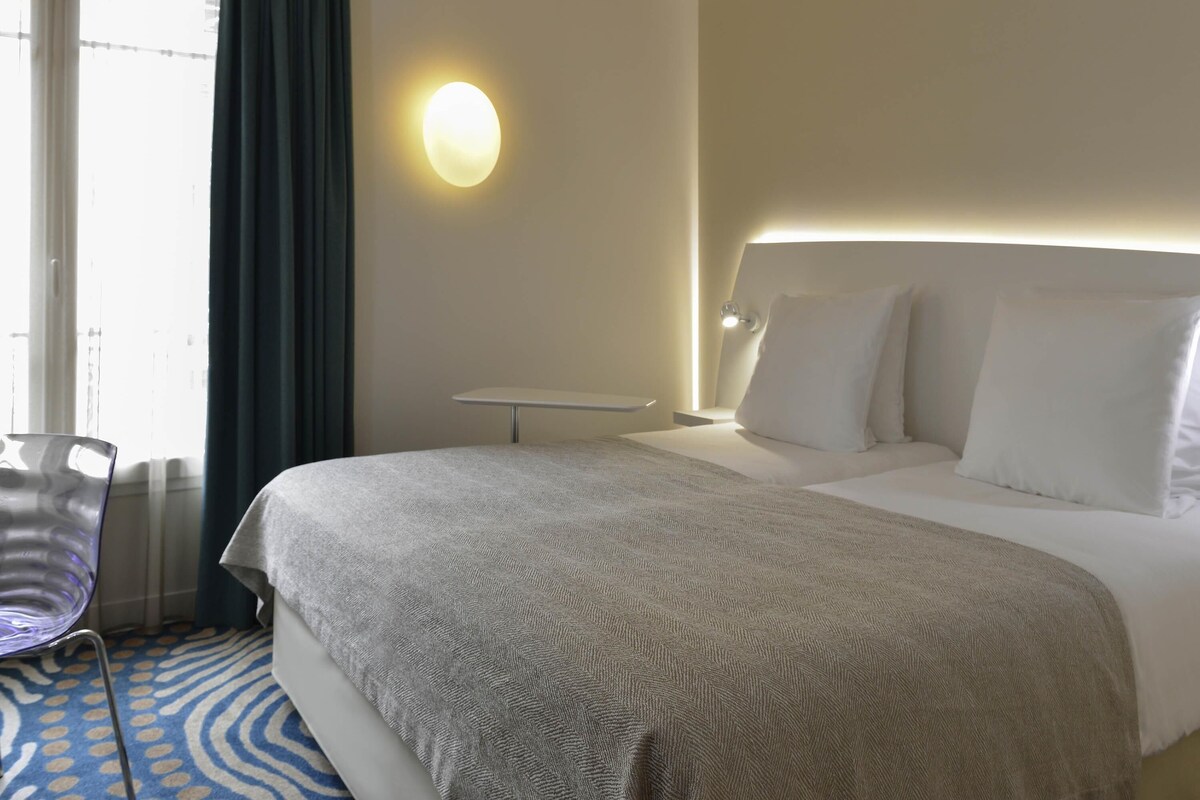 A hotel room with a bed, bedside table, lamp, chair, and a wall-mounted light fixture.