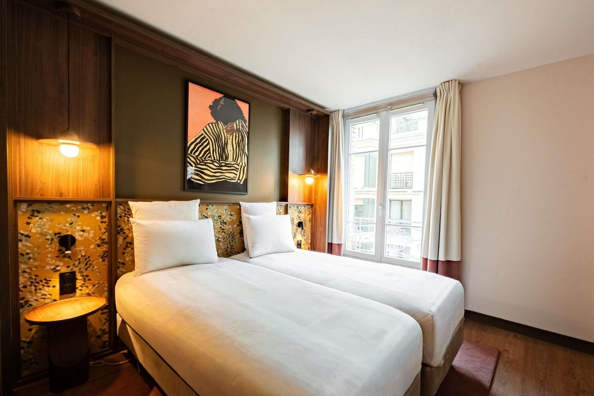 A hotel room with two beds, a large window with curtains, a wall-mounted artwork, and a small table with a lamp.