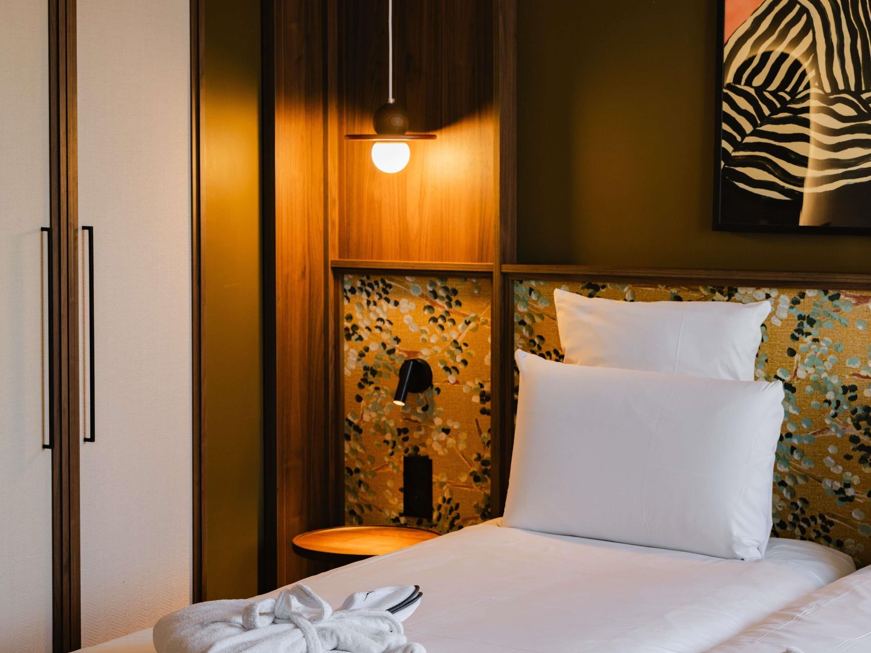 A hotel room with a bed, a nightstand, a lamp, and a wall-mounted artwork.