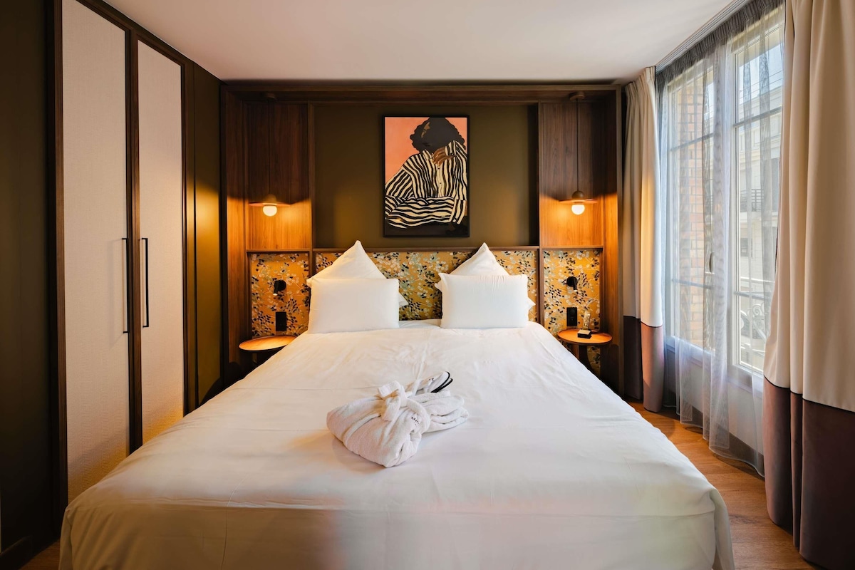 Une chambre d’hôtel avec un grand lit, deux tables de chevet, une œuvre d’art accrochée au mur et une fenêtre avec des rideaux.