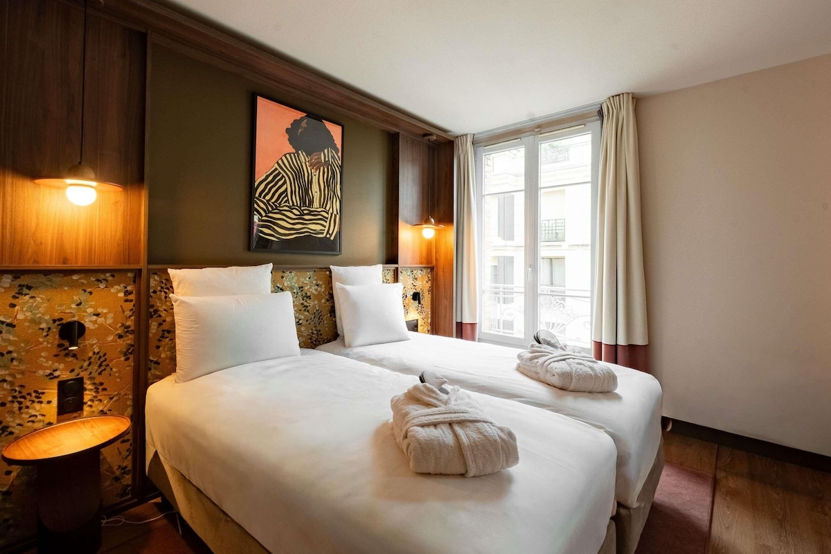 Une chambre d’hôtel avec deux lits, une table de chevet, une œuvre d’art accrochée au mur et une fenêtre avec des rideaux.