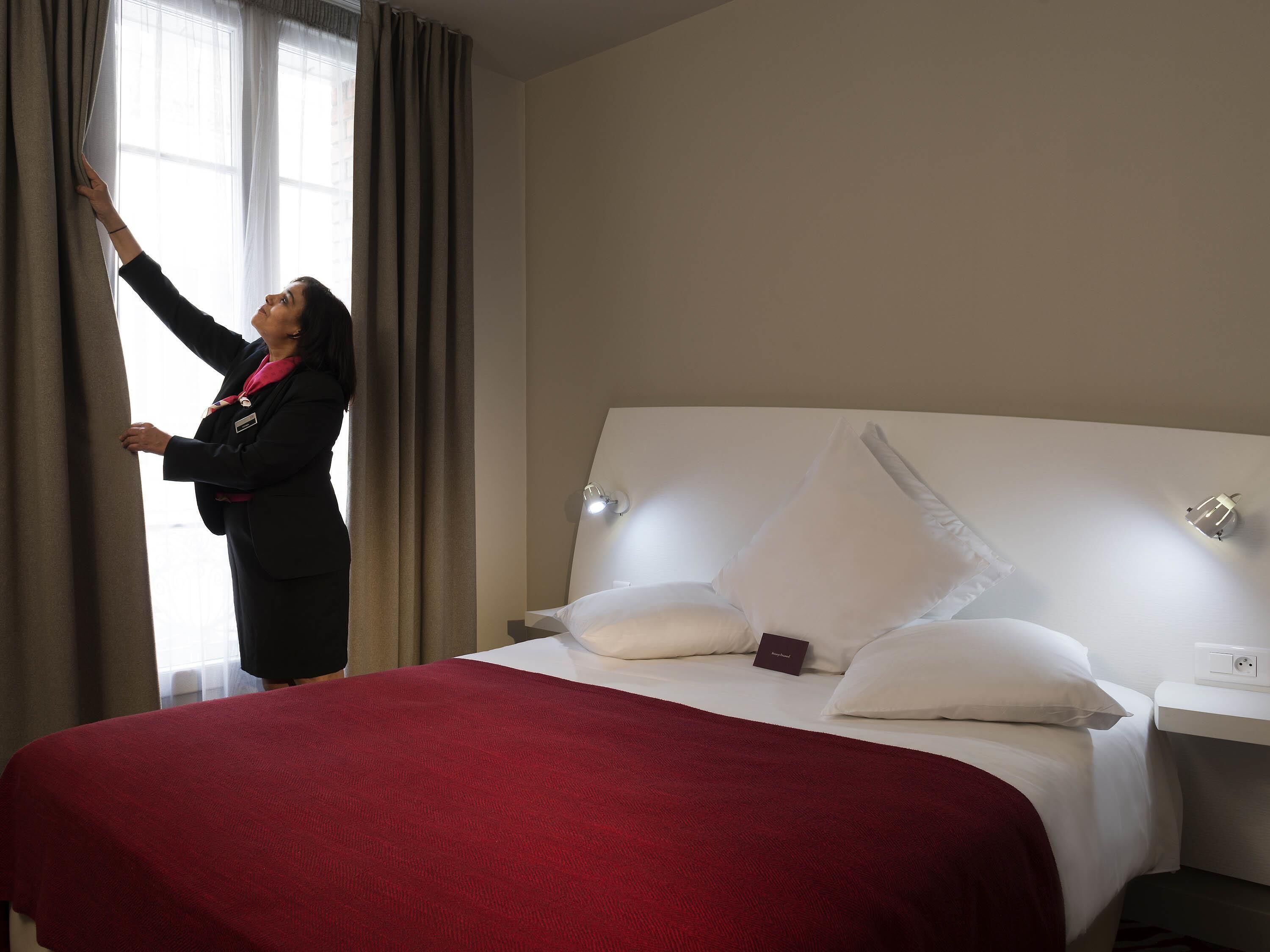 A hotel room with a bed, a bedside table, a lamp, and a window with curtains.