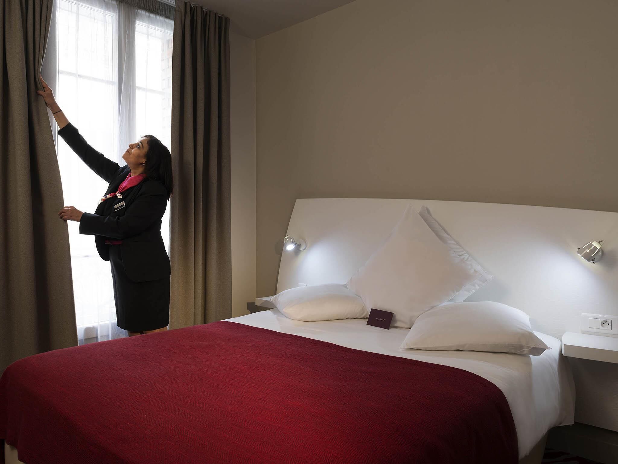 Una habitación de hotel con cama, mesita de noche, lámpara y ventana con cortinas.