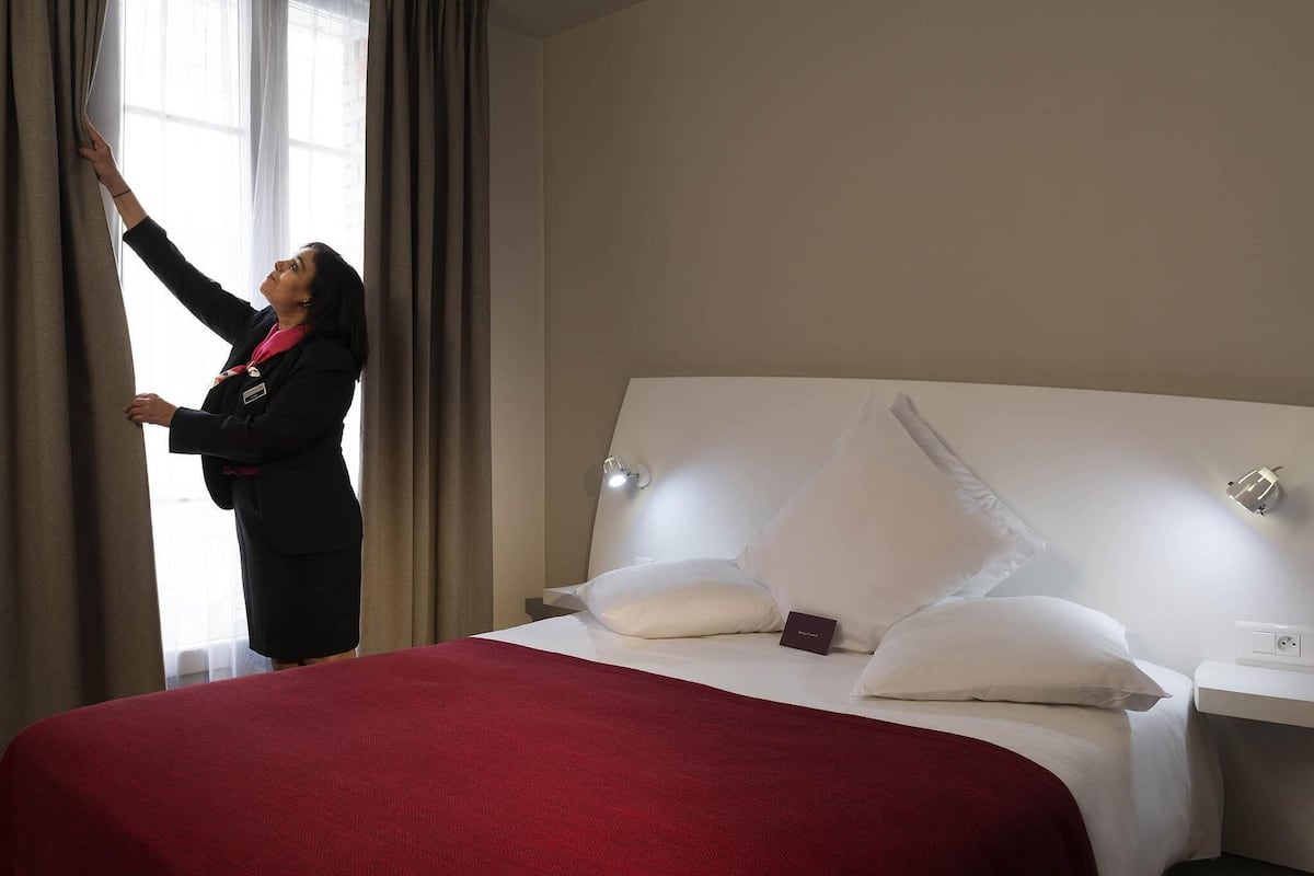Una habitación de hotel con cama, mesita de noche, lámpara y ventana con cortinas.